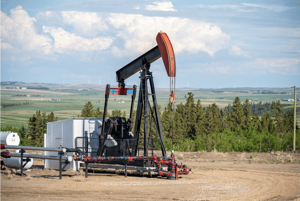 Oil pumpjack operating at a well site with pipelines, equipment, and a rural landscape in the background.